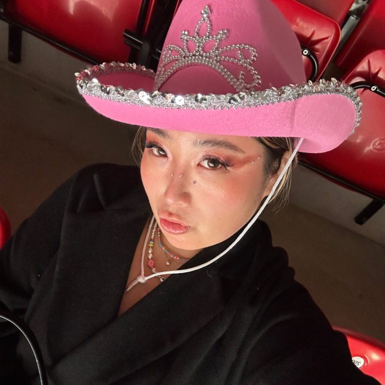 Woman wearing a pink cowboy hat with rhinestones, seated in a theatre-like environment.