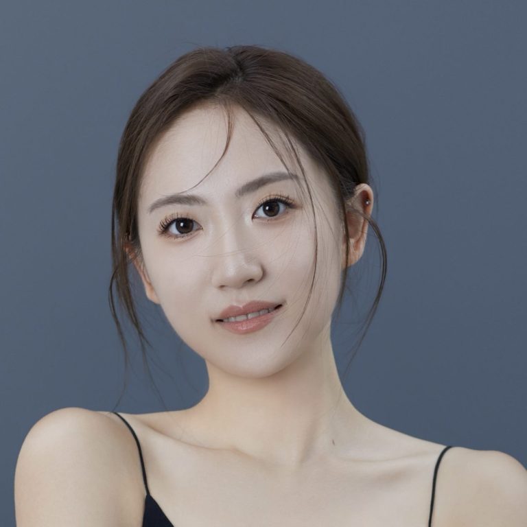 A young woman with long dark hair wearing a black top, smiling softly against a grey background.